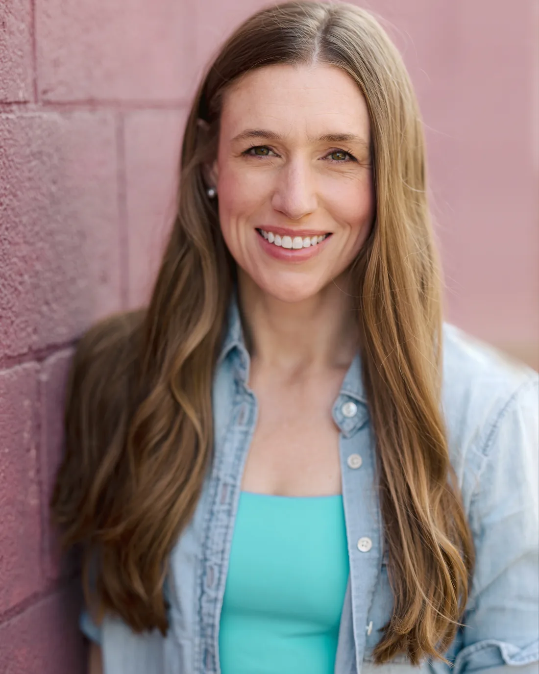 megan, who makes every hello paint kit by hand, smiling in a denim shirt against a pink brick wall